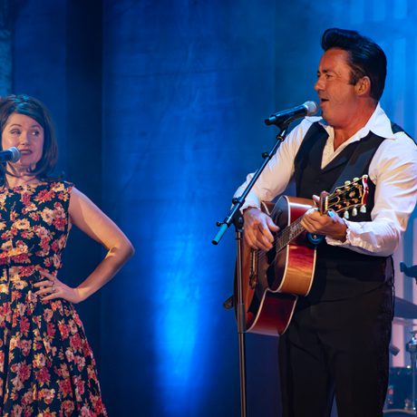 A man in a white shirt and black waistcoat playing a guitar. He is singing with a woman with brown shoulder-length hair in a black and red floral dress.