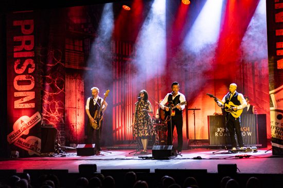 A band and singers on stage, lit in red lighting. They are playing guitar, keyboard, and drums.