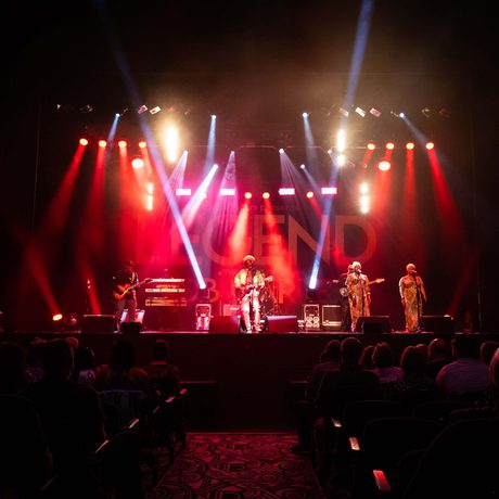 Multiple performers singing and playing guitars on a stage. The stage is lit with red spotlights.