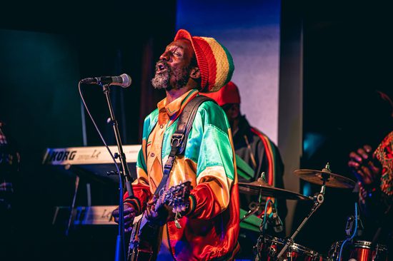 A black man wearing a hat and jacket that are red, yellow and green. He is singing into a microphone and playing the guitar.