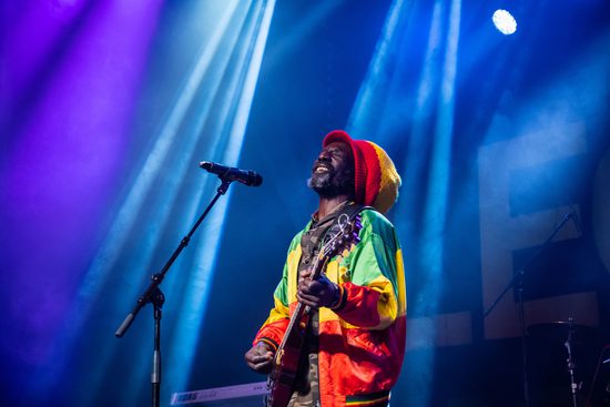 A black man wearing a hat and jacket that are red, yellow and green. He is smiling and playing the guitar under blue lighting.