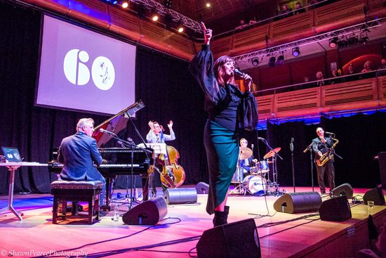 A stage lit in pink with various performers playing instruments, including piano, double bass, drums and saxophone. There is a lady in a black top and teal skirt singing into a microphone.