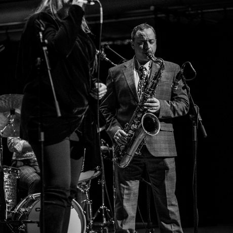 Black and white image of a woman singing into a microphone. There is a man behind her playing the saxophone.