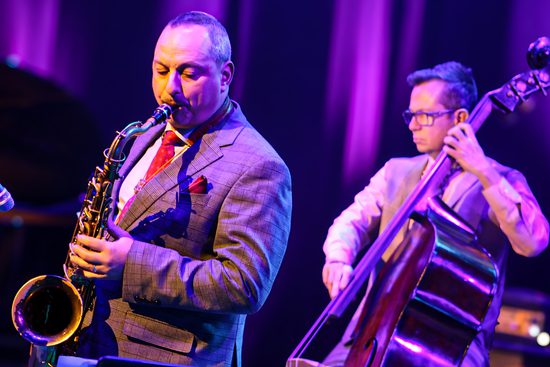 A man in a grey suit and red tie playing the saxophone. A man behind him plays the double bass. The lighting creates a pink hue.