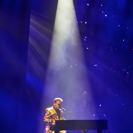 A man waring a gold suit plays piano under a single spotlight.