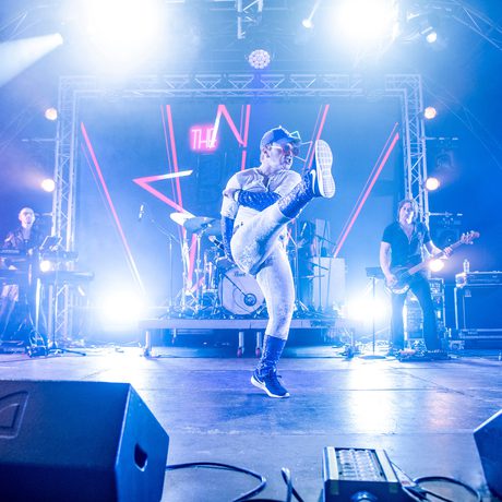 A man wearing white trousers, a white shirt, and a blue baseball cap dances across a stage.