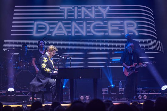 A white man wearing glasses and a black jacket with gold embellishments plays piano on a stage. The sign in the back says Tiny Dancer.