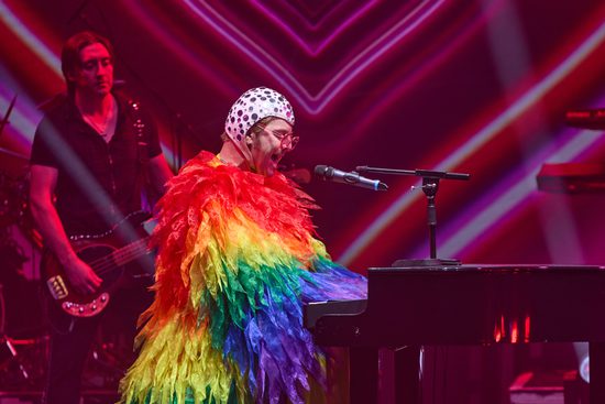A man wearing a large, rainbow, feathered outfit and a white cap with jewels on it plays the piano.