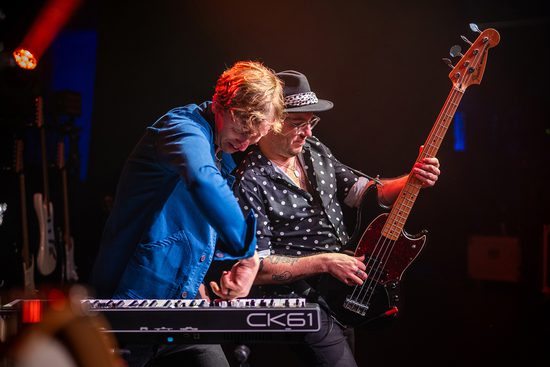 Man in blue shirt plays the keyboard and a man in a polka dot shirt and fedora hat plays a bass guitar. They lean in towards each other.
