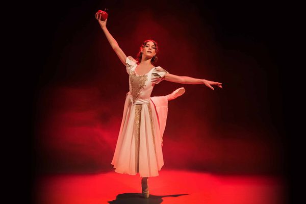 A white, brunette woman wearing a gold and white dress dances en pointe, holding a red apple in the air.