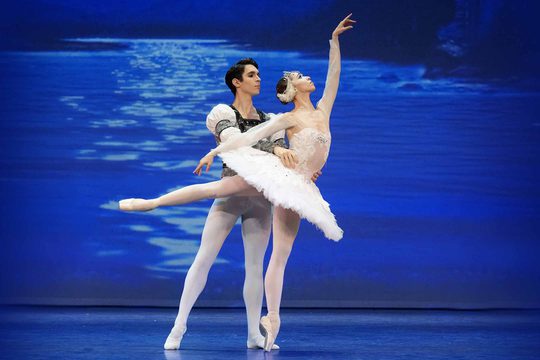 A male ballet dancer wearing a white shirt and tights and a female ballet dancer in a white tutu dance in front of a blue background depicting water.