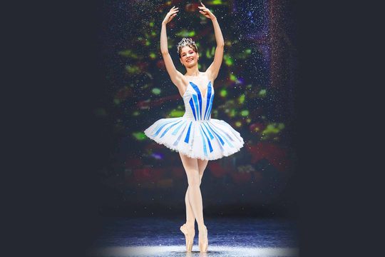 A ballerina in a blue and white tutu with both hands above her head.