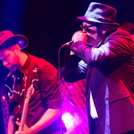 A man in a hat and sunglasses plays a harmonica into a microphone. He is lit by red lighting.