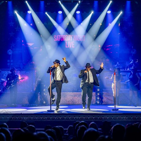 Two men in black suits, white shirts, black ties, hats and sunglasses sing and dance on a stage.
