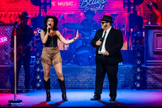 A woman in a black corset top, brown shorts and a cowboy hat sings with a man wearing a black suit, white shirt, hat and sunglasses.