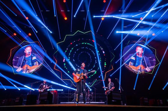 A man on a stage singing whilst playing an acoustic guitar. He is surrounded by blue lasers and strobe lights.