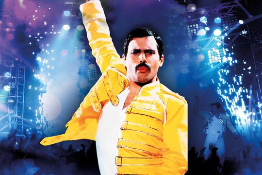 A man in a yellow jacket and white top underneath stands with one arm in the air. He has dark hair and a moustache.