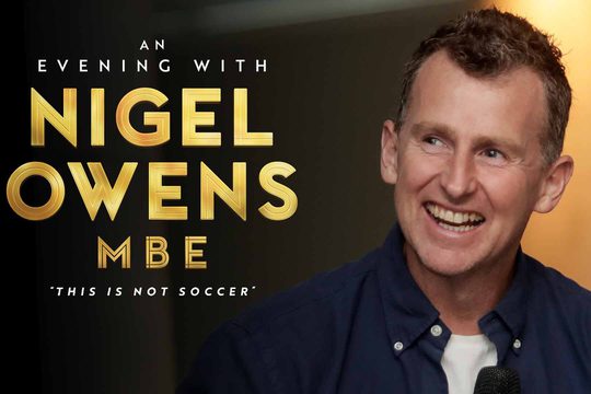 A white, brunette man wearing a navy blue shirt smiles. Gold text: An evening with Nigel Owens MBE. "This is not soccer".