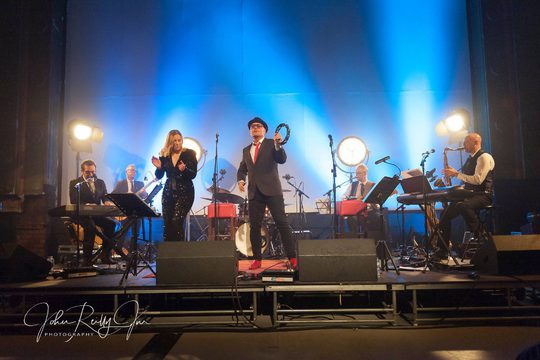 A collection of musicians on a stage playing various instruments. The stage is lit blue.