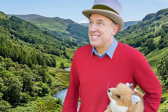 A man in a red jumper and a top hat stands in front of a lush green valley.