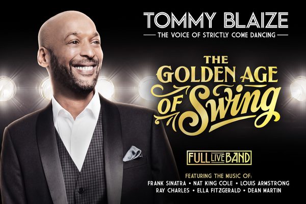 A bald man with a beard smiles in front of stage lights. He is wearing a black suit.