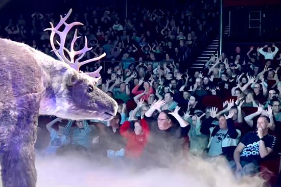 A realistic reindeer puppet looking out into an audience. All of the audience members have their hands on their head to create reindeer antlers.