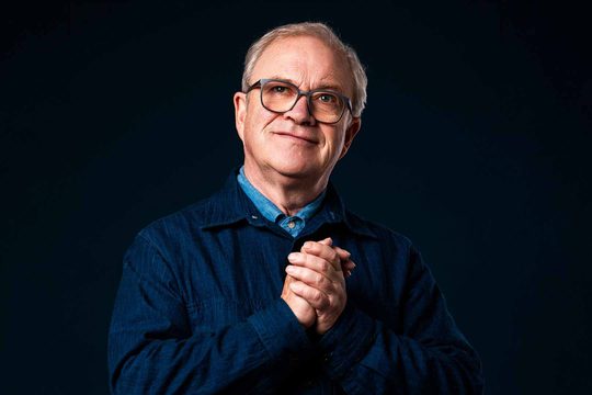 A man standing in front of a black background. He is wearing a dark blue jacket, light blue shirt, and glasses. His hands are clasped together in front of his chest.