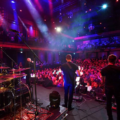 A band playing instruments, wearing all black. The image is taken from behind the band, so you can see a full audience watching them.