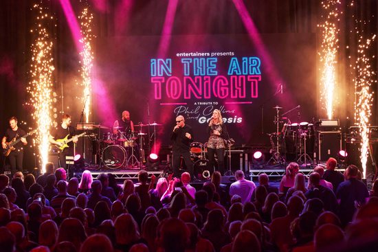 A bald, white man wearing sunglasses and black clothes and a white, blonde woman wearing sparkly clothes sing on a stage to an audience. There are fireworks around them.