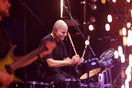 A bald, white man wearing all black plays the drums, with fireworks around him.