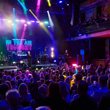 A crowd watching a band perform. The room is lit with spotlights in various colours, including pink, blue, yellow, and orange.