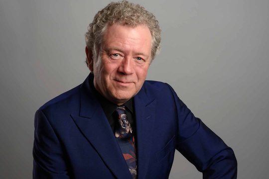 An older, white man with grey curly hair looks straight at the camera and smiles. He is wearing a dark blue suit and floral tie.