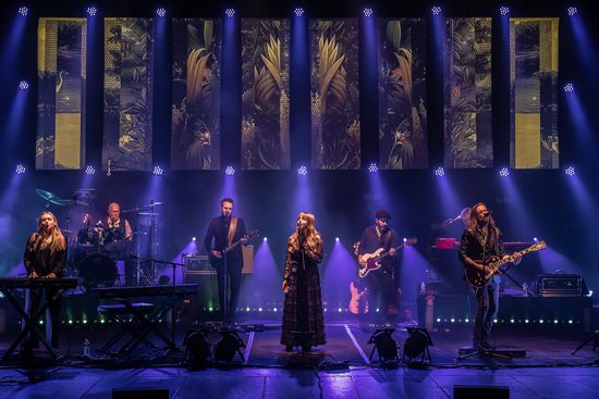 A band wearing black perform on a stage lit in purple. Panels on the back of the stage show floral and leafy patterns.