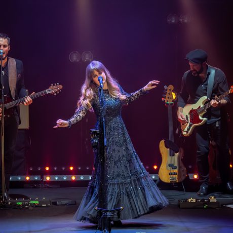A woman in a black, sparkly, long dress sings into a microphone. Two men wearing black play guitar behind her.