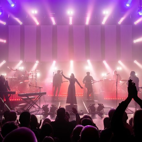 Silhouettes of a band playing on a stage. They are lit in pink and purple. In the foreground an audience gives them a standing ovation.