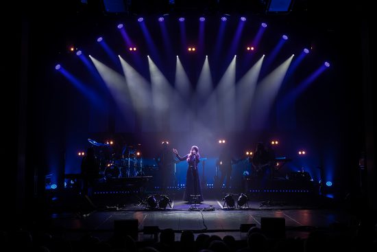 A band playing on a stage with a female singer in the centre. The stage is dark apart from multiple spotlights all pointing on her.