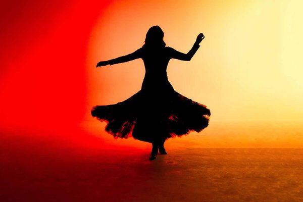 Black silhouette of a woman wearing a long dress with a big skirt. She is dancing in a red and yellow sunrise.