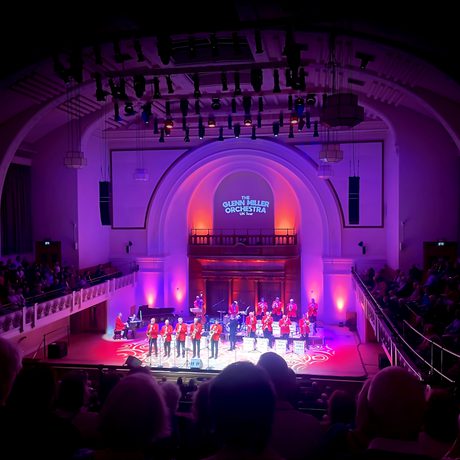 A big band playing on a stage to a large audience. They are lit in pink and are wearing red jackets.