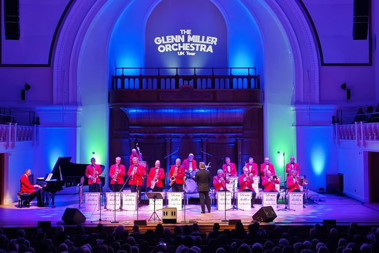 A big band playing instruments on a stage lit in blue. They are all wearing red jackets.