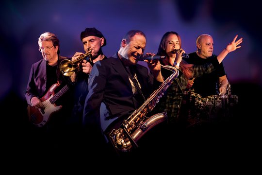 Five musicians against a purple background. They are playing electric guitar, trumpet, saxophone, drums, and singing.