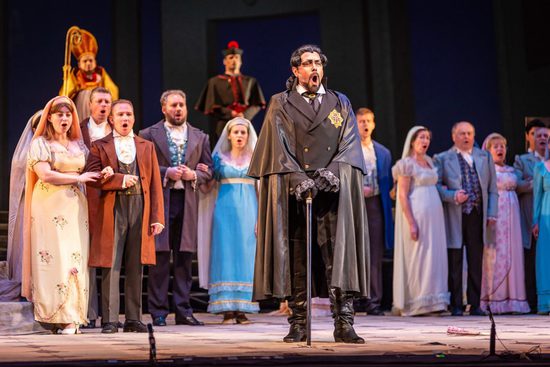 The cast stand in a line all facing the same direction and singing. In front is a man wearing a black wig, jacket and cape, and holding a black walking stick.