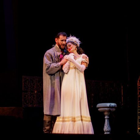A woman in a white dress and a feathered headdress and a man in a grey jacket embrace each other.