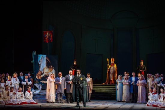 A large cast all stand on the stage. The scene is in a church, with choristers and nuns either side, and a couple getting married in the middle.