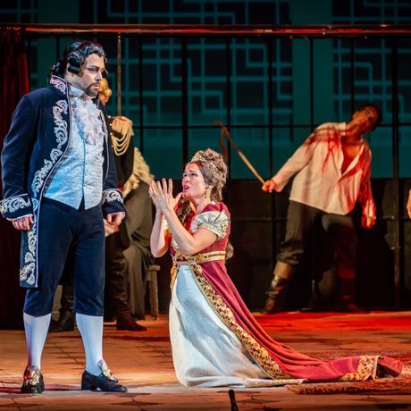 A woman in a red and white dress with a gold crown kneels on the floor in front of a man in a black wig and ornate navy blue jacket.