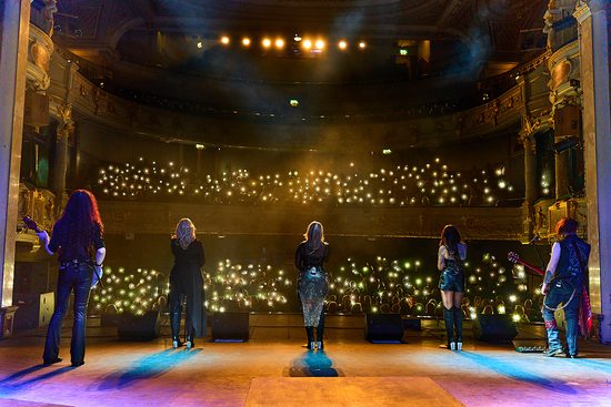 Three female singers and two guitarists sing on a stage. The picture is taken from behind them, and you can see the audience holding up lights.