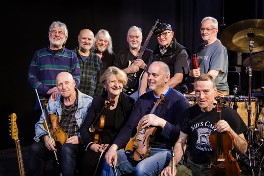 A group of ten musicians holding musical instruments including violins and a bass guitar. They are all smiling.