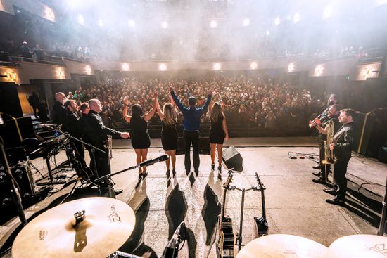 Picture taken from the back of a stage showing a band bowing to a standing ovation from a sold-out audience.