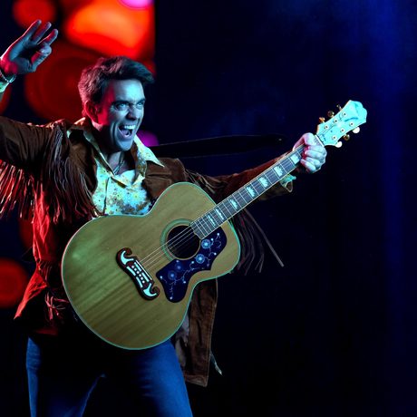 A man wearing blue jeans and a brown fringed jacket plays a brown acoustic guitar. He has one arm in the air and his mouth is open.