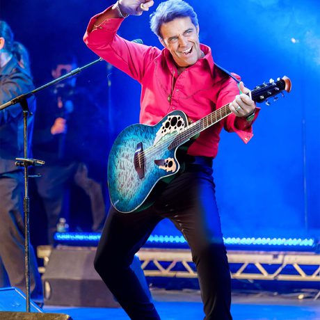A man in black trousers and a red shirt plays a blue guitar. He is lit in blue lighting.
