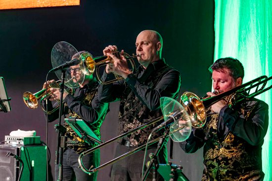 Three men wearing black playing brass instruments. They are lit with green lighting.